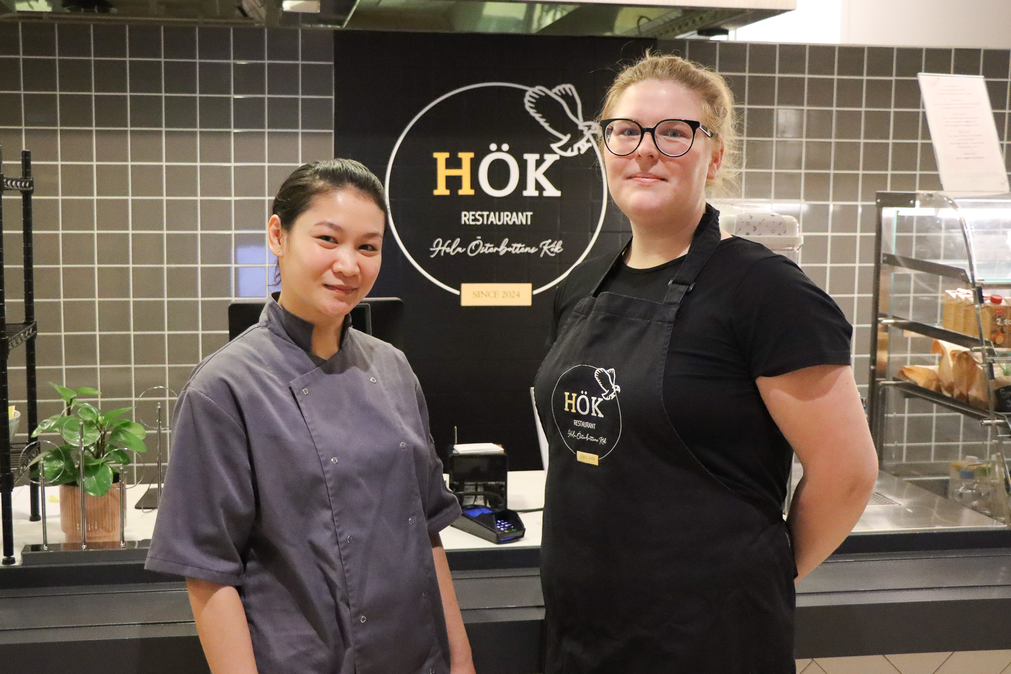 HÖK-ravintolan omistaja ja oppisopimusopiskelija seisovat palvelutiskin edessä ja hymyilevät kameralle.